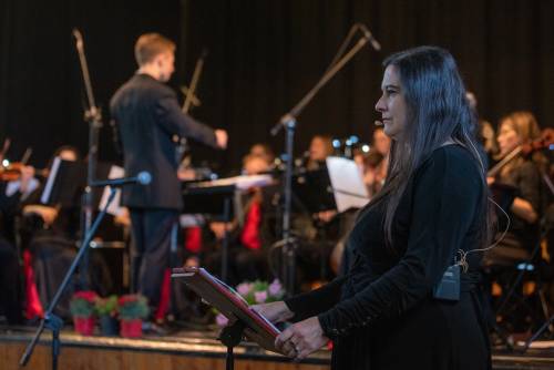 Zdjęcie przedstawia prowadzącą koncert z orkiestrą w tle.